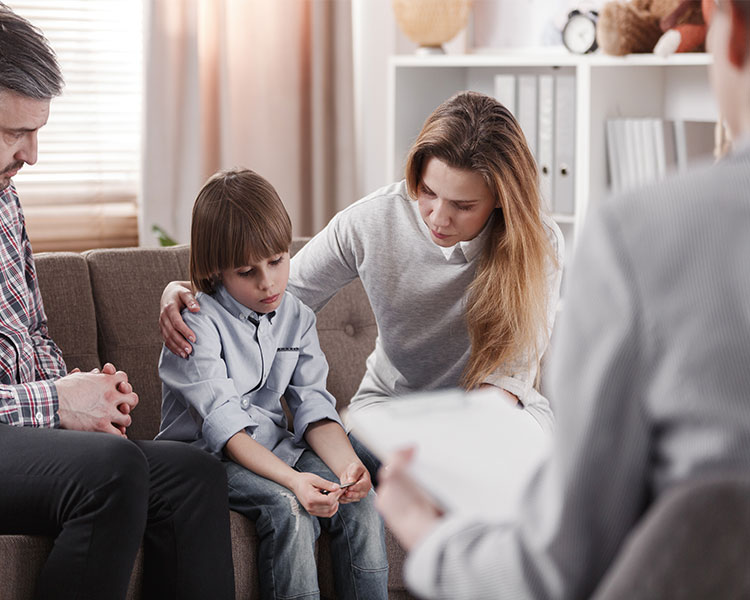 A child wearing a sad expression while sitting on a couch with their parents across from a therapist