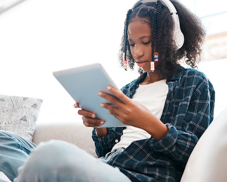 Image of a teen sitting on a couch and wearing headphones while using a tablet